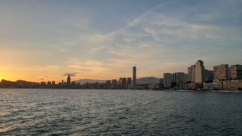 Sea by buildings in city against sky during sunset