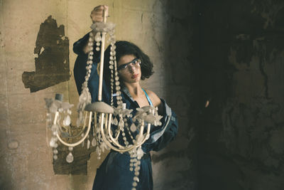 Portrait of young woman holding chandelier while standing against wall