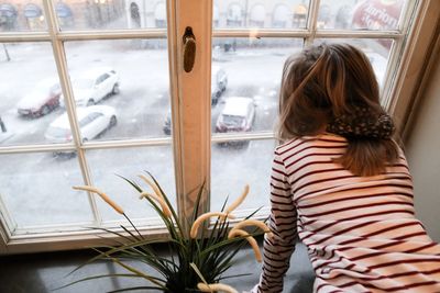 Rear view of girl looking at snow covered cars on road through window