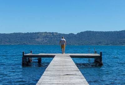 Rear view of man standing on pier over lake against clear blue sky