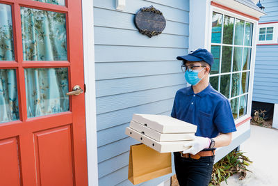A male delivery man holds a box of goods and food for a customer in front of the house.