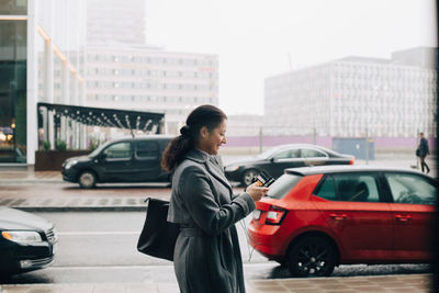 Side view of businesswoman using smart phone while walking on sidewalk against sky in city