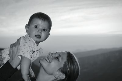 Close-up of happy mother carrying baby girl against sky