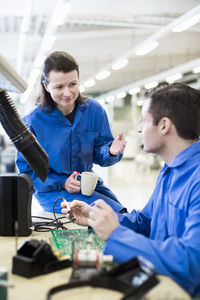 Female technician gesturing while looking at colleague working on circuit board at desk