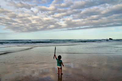 Rear view of toddler holding  branch on deserted beach against sky