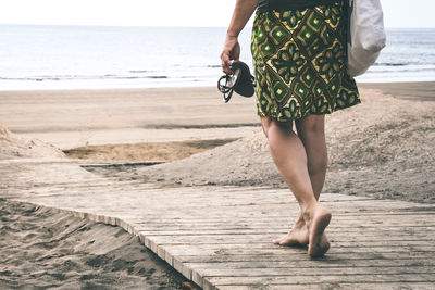 Low section of woman walking on boardwalk leading towards beach