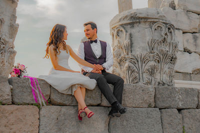 Smiling bride with groom sitting on stone wall at old ruin