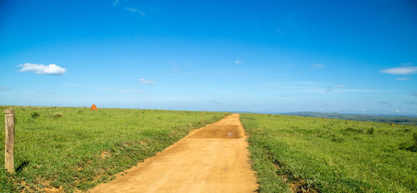 Dirt road amidst field against blue sky
