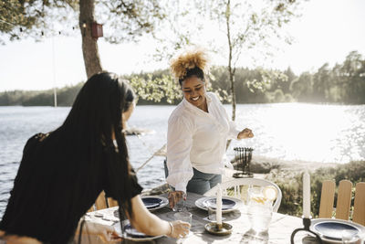 Smiling woman setting table with female friend during dinner party on sunny day