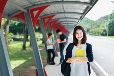 Portrait of happy young woman holding book while friends discussing in background at college campus