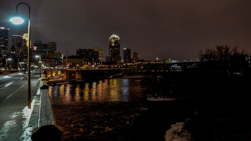 Illuminated street light on road by river against buildings at dusk