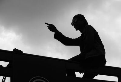 Low angle view of silhouette man pointing while sitting on metal against sky