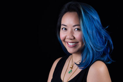 Close-up portrait of happy woman with blue hair standing against black background