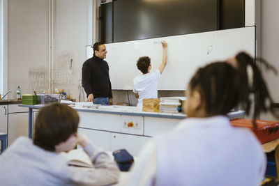 Smiling teacher standing with schoolboy writing on whiteboard in classroom