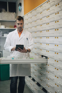 Mature male pharmacist holding digital tablet standing by drawer at store