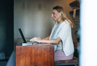Young adult blonde woman with long hair working on laptop and using mobile phone on kitchen at home