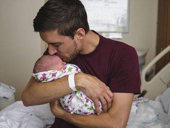 Father kissing newborn son while sitting on bed at hospital