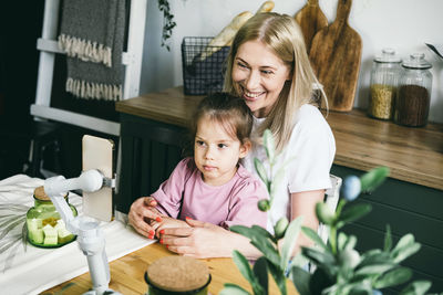 Happy mother and daughter waving hands while looking at webcam in smartphone on tripod for videocall