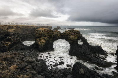 Wave splashing on rock formation against cloudy sky