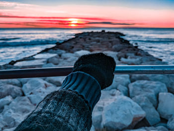 Low section of person on rock at sea shore during sunset