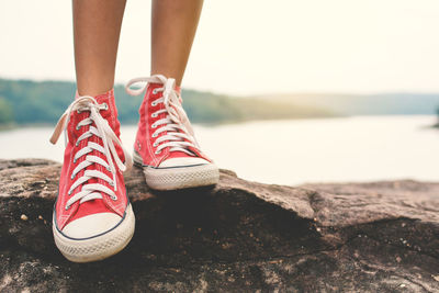 Low section of woman wearing red shoes standing on rocks by lake