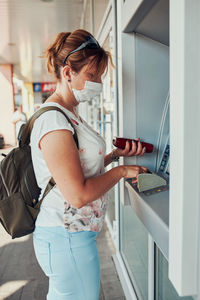 Woman withdrawing money from atm machine using debit or credit card standing outdoors in the street