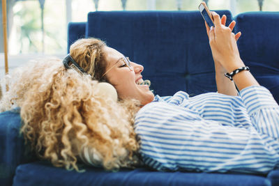 Side view of cheerful woman using smart phone while lying on sofa at home