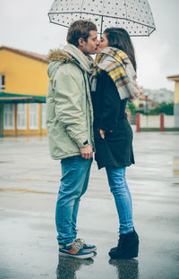 Side view of couple wearing warm clothing while kissing under umbrella in city