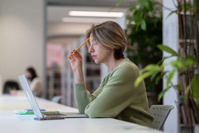 Concentrated female mature student studying online on laptop in modern library surrounded by green