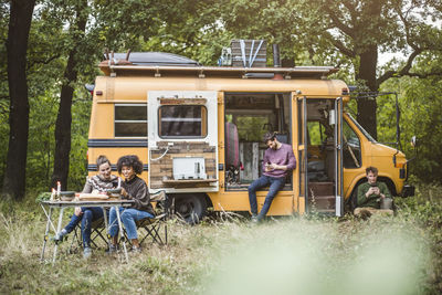 Smiling female friends sitting at table while men using smart phones during camping in forest