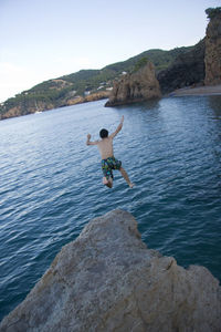 Man jumping on rock by sea against clear sky