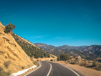 Empty road by mountains against clear blue sky