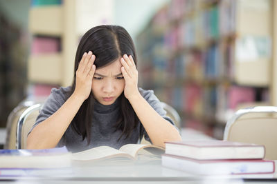 Tensed young woman reading book while sitting at table in library