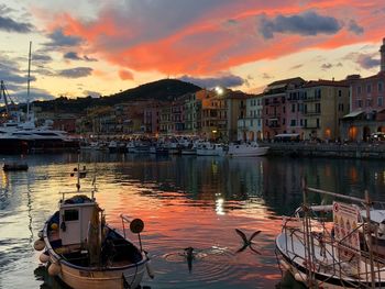 Boats moored at harbor by buildings against sky during sunset