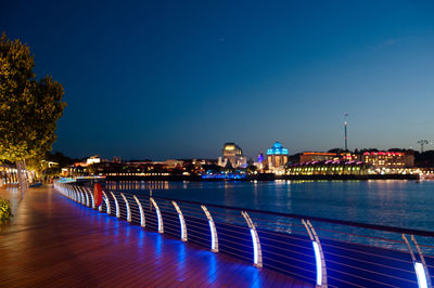 Illuminated city by river against clear blue sky at night