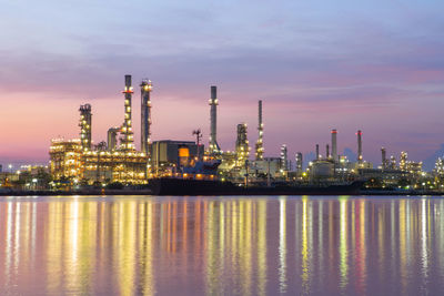 Illuminated factory by river against sky at dusk