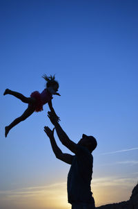 Low angle view of silhouette man catching daughter against blue sky during sunset