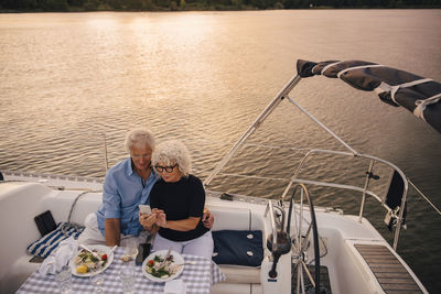 High angle view of senior couple taking selfie through mobile phone in sailboat during sunset