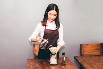 Young woman pouring coffee in filter on table against wall