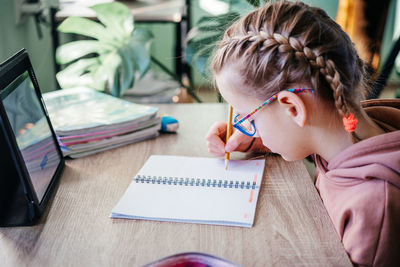 Primary school girl in eyeglasses writing in her notebook while having online lesson during covid