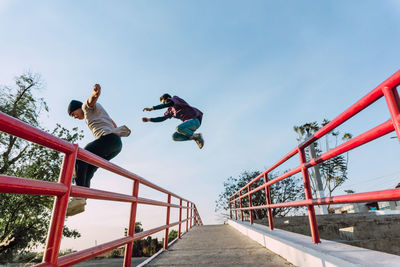 Low angle side view of fearless male friends jumping above metal railing in city while performing parkour stunt on sunny day