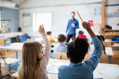 Rear view of students sitting with hands raised in classroom