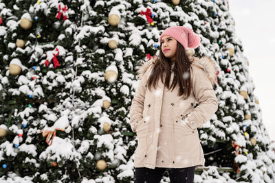 Happy woman in warm winter clothes standing by the big christmas tree outdoors, snow falling