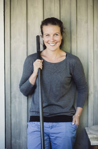 Portrait of happy woman holding shovel while standing against wall