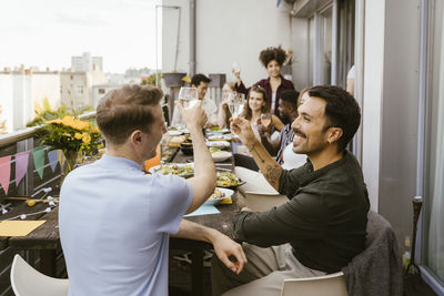 Happy men toasting drinks with group of friends sitting at dining table in balcony