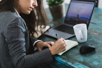 Side view of cropped unrecognizable focused young female entrepreneur in formal wear sitting at table with laptop and cup of coffee and writing information in notebook while planning working day at workplace at home