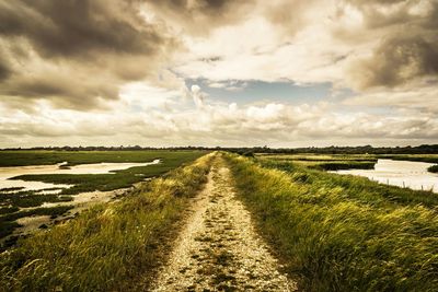 Road passing through field against cloudy sky