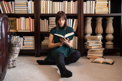 Young woman reading book while sitting against bookshelf in library