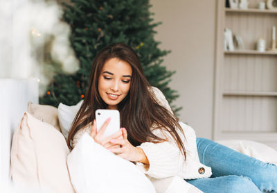 Young asian woman in cozy white knitted sweater using mobile on bed in room with christmas tree