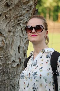Portrait of young woman wearing sunglasses standing on tree trunk